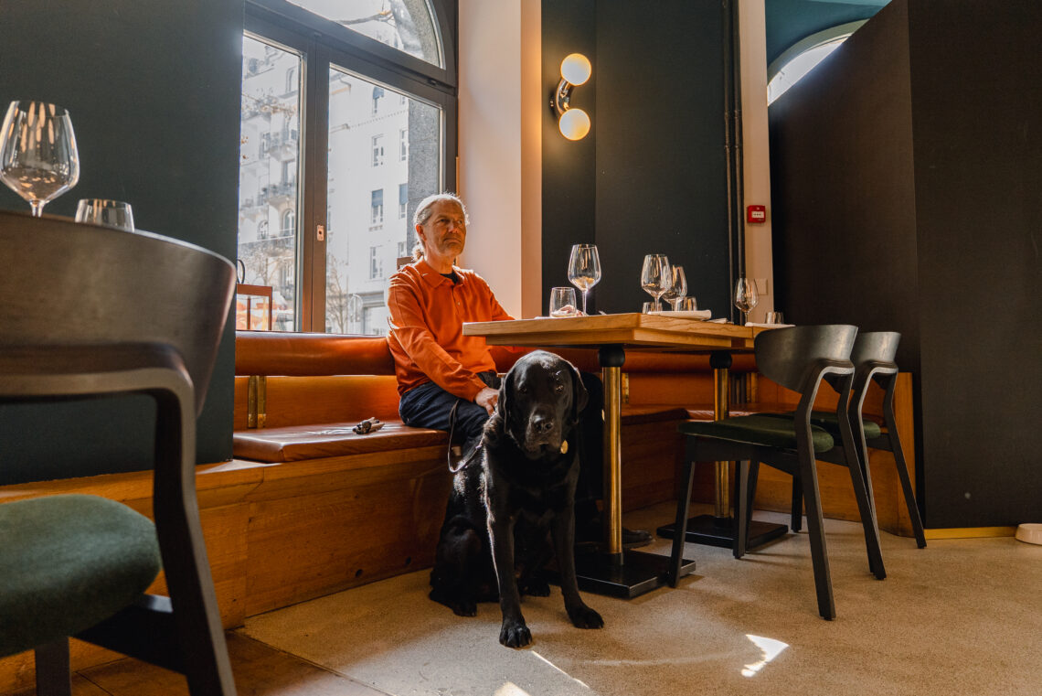 Eine blinde Person sitzt an einem Tisch im Restaurant. Hinter der Person scheint die Sonne durch ein hohes Fenster. Unter dem Tisch sitzt ein schwarzer Blindenführhund und schaut müde in die Kamera. 