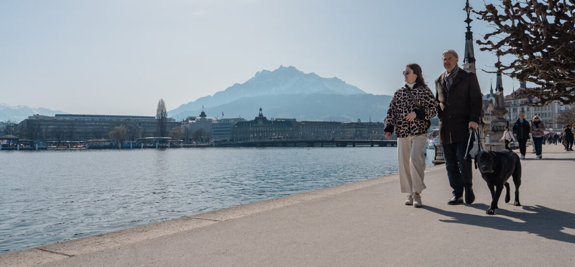 Eine Frau und ein Herr gehen dem See entlang. Der Mann ist bei der Frau eingehakt. In der anderen Hand hat der Mann einen Blindenstock und die Leine des schwarzen Blindenhundes, der gemütlich neben den zwei Personen hergeht. Die Sonne scheint, das Wasser des Sees ist türkisblau und im Hintergrund sind verschneite Berge.
