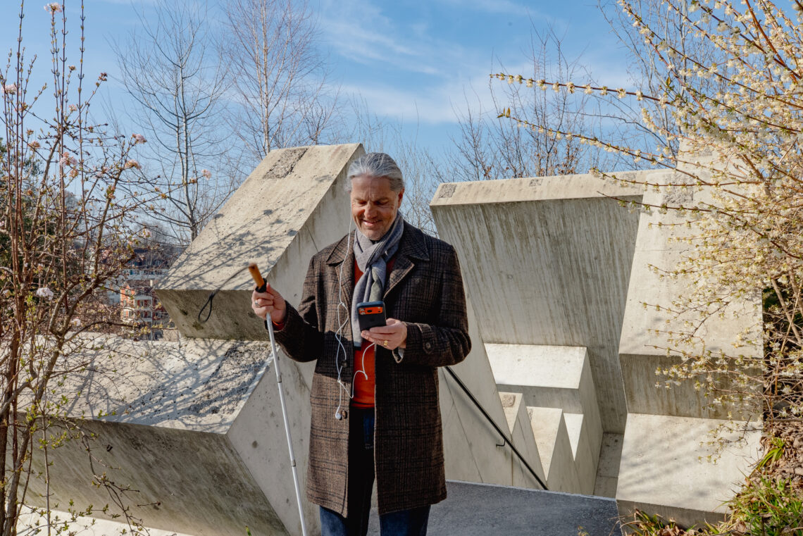 Eine blinde Person erkundet mit einem Audio-Guide ein Museum. Mit hellen Kabelkopfhörern im Ohr, dem Handy in der einen Hand und dem Blindengehstock in der anderen Hand steht die Person vo einem Betongebäude im Gletschergarten Luzern. Die Sonne scheint und der Himmel im Hintergrund ist blau. 