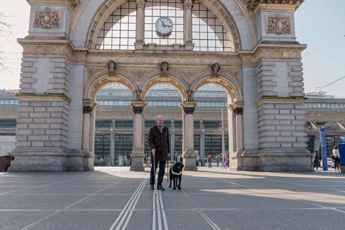 Eine bline Person läuft in Begleitung eines schwarzen Blindenhundes entlang der Blindenführungslinien vor dem Torbogen in Luzern. Die Sonne scheint durch den Torbogen.