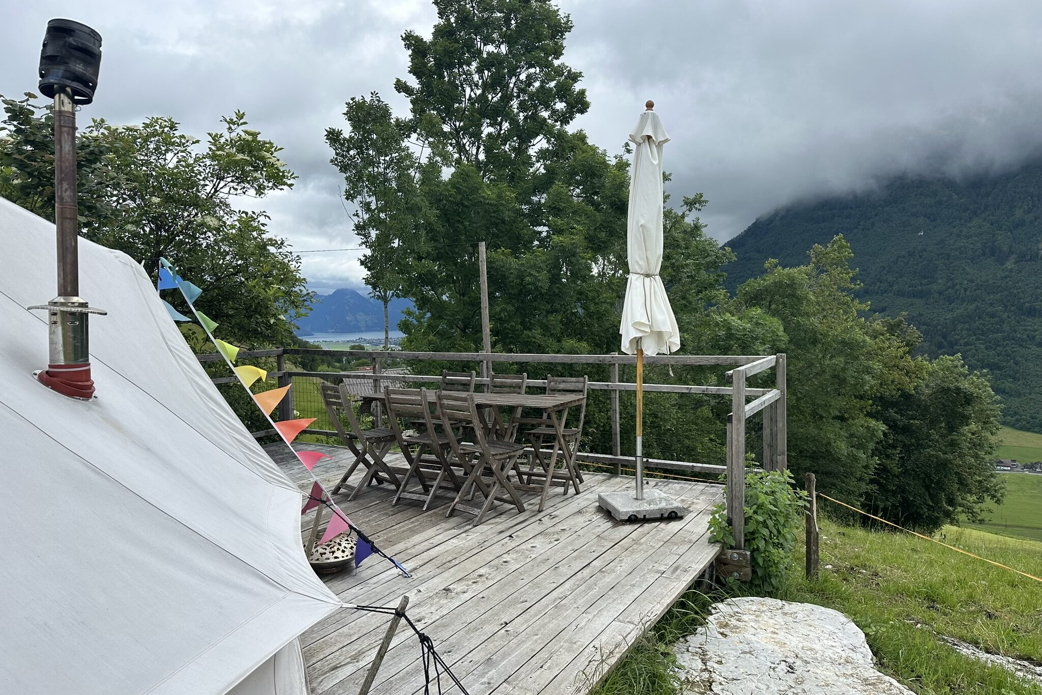 Bellzelt auf Holzplattform mit Gartentisch, Stühlen und geschlossenem Sonnenschirm.